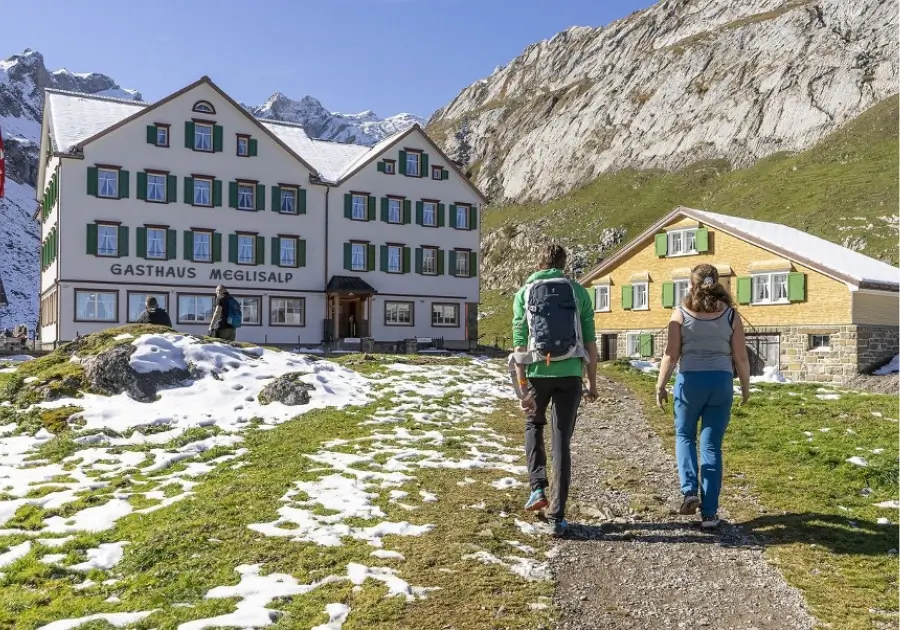 Two people hiking on a path toward a large guesthouse and a smaller house in a snowy mountain valley, with clear skies and rocky peaks in the background.
